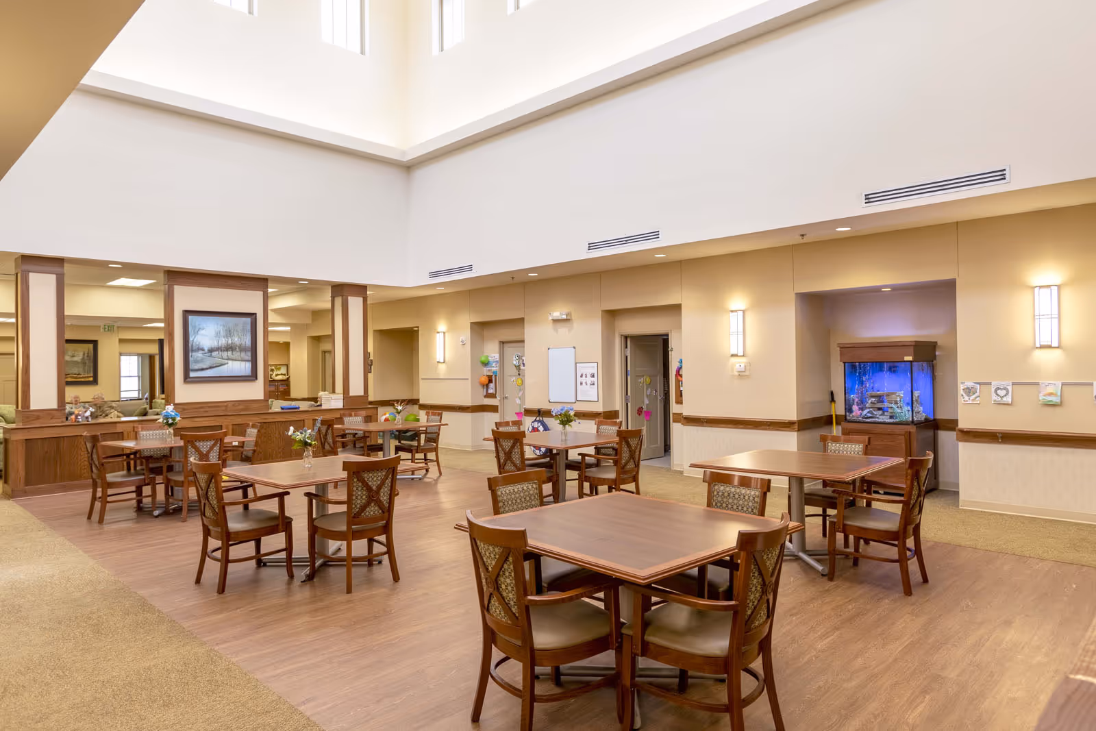 A spacious and well-lit dining area in a senior living facility with several wooden tables and chairs arranged neatly on a wood floor. The room features high ceilings with large windows near the top, beige walls with wooden trim, wall-mounted lights, and a large fish tank in a recessed wall area. There are paintings on the walls and some decorations on the doors in the background.
