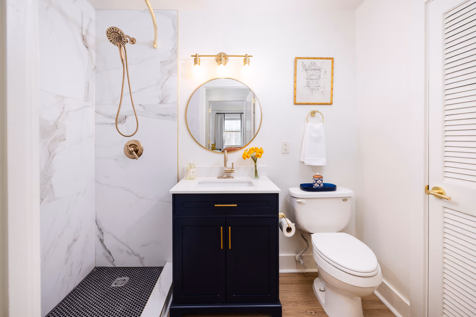 A modern bathroom featuring a walk-in shower with marble-patterned walls and a black hexagonal tile floor. There is a dark blue vanity with a white countertop, a round mirror above it, and a gold light fixture. A white toilet is next to the vanity with a towel ring holding a white towel and a decorative tray on top of the toilet tank. The bathroom has light-colored walls and a wooden floor.