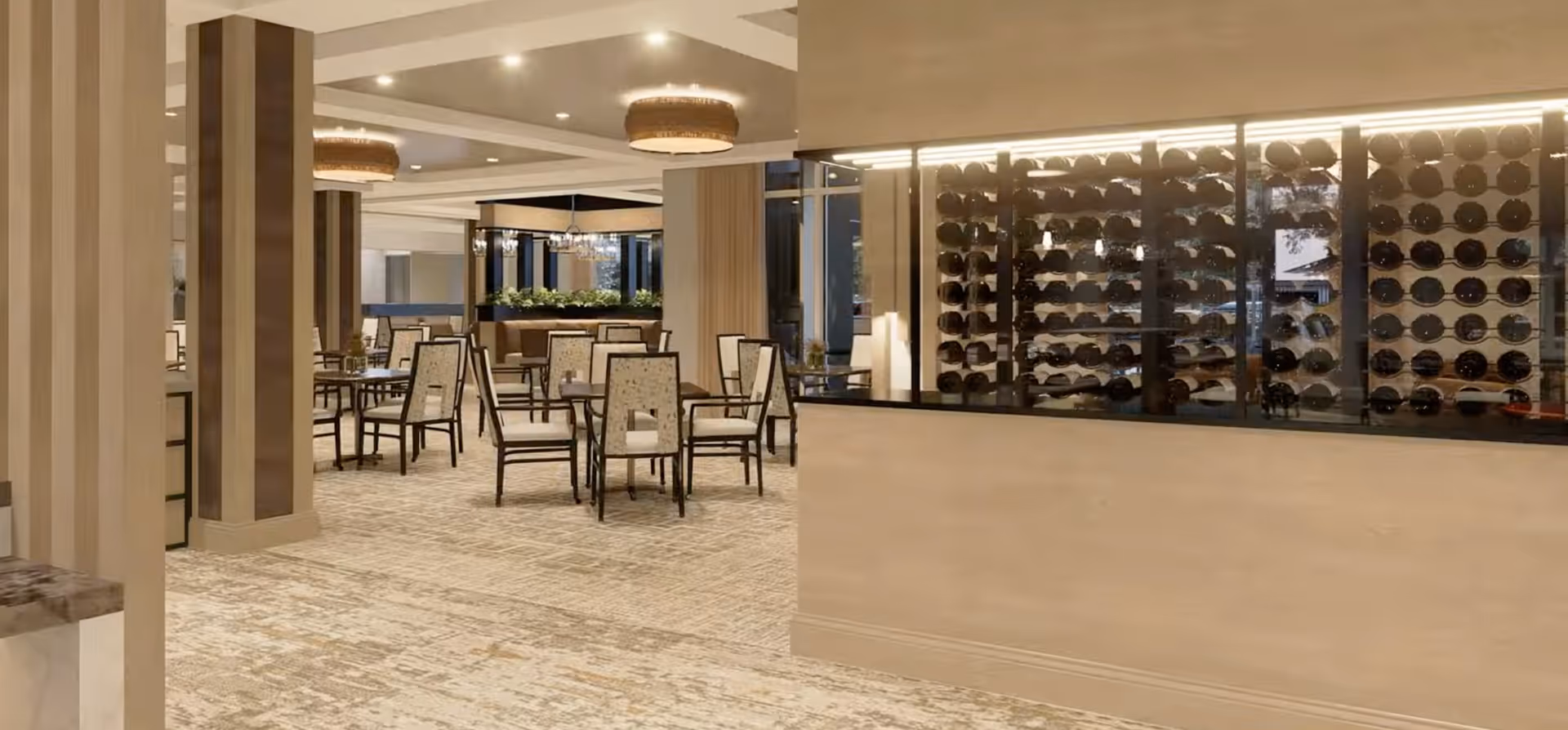 Interior view of a dining area with multiple tables and chairs arranged neatly. The room features beige walls, carpeted flooring, and modern ceiling lights. On the right side, there is a glass-enclosed wine rack displaying numerous wine bottles.