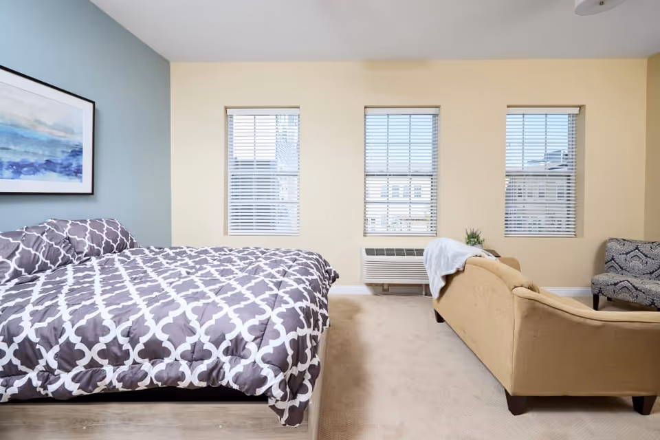 A bright room with a bed featuring a gray and white patterned comforter on the left side, three windows with blinds in the center, a beige couch with a white blanket draped over the back on the right, and a patterned armchair in the corner. A framed abstract painting with blue tones hangs on the left wall.
