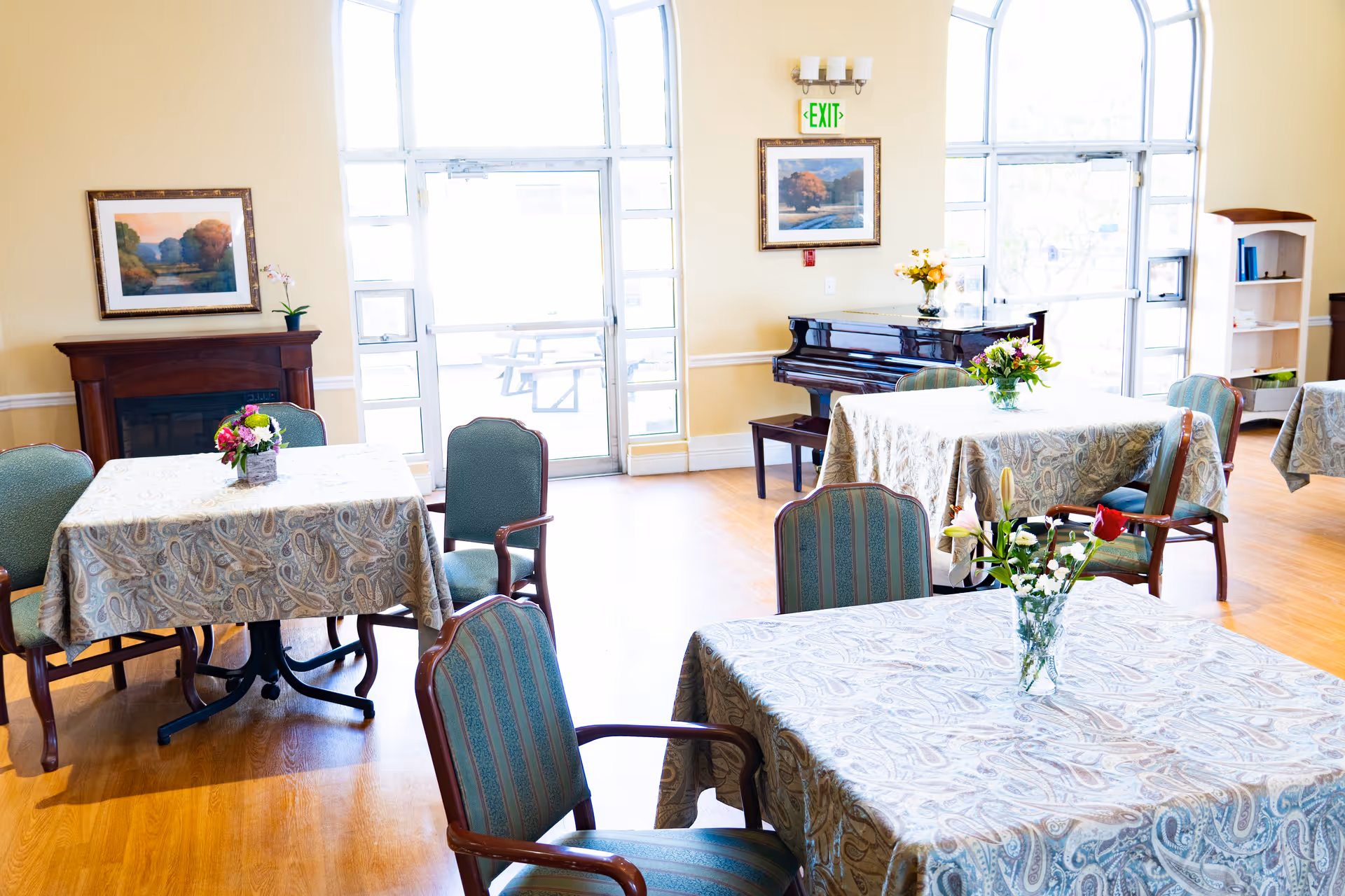 A bright dining room with several tables covered in patterned tablecloths, each adorned with a vase of flowers. The room features large windows and glass doors letting in natural light, a piano against the wall, framed landscape paintings, and a wooden floor.