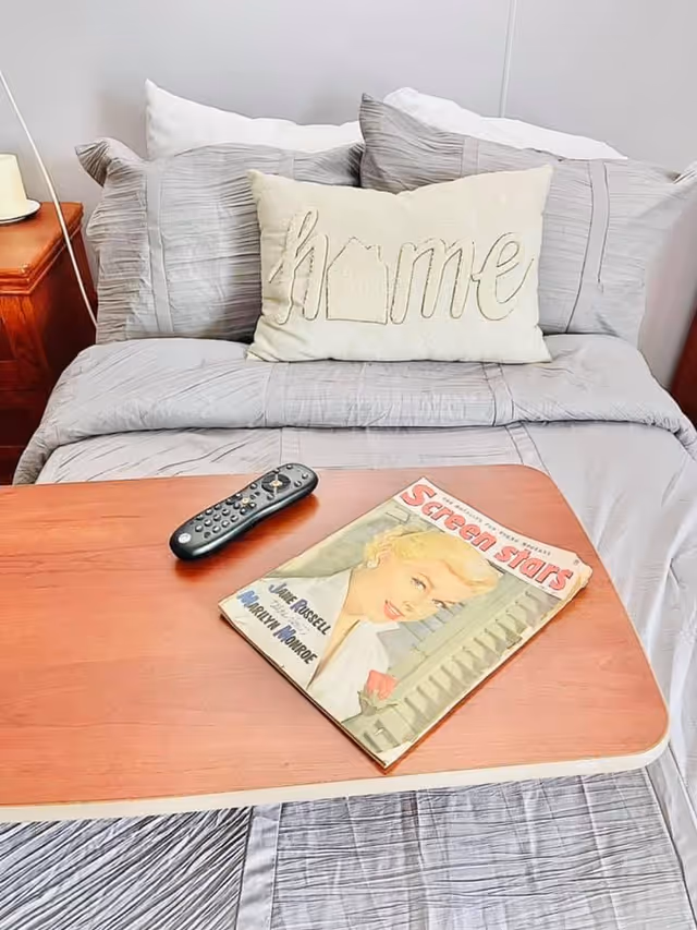 A neatly made bed with gray striped bedding and a decorative pillow with the word 'home' embroidered on it. On the bed is a wooden overbed table holding a TV remote and a vintage magazine titled 'Screen Stars' featuring a portrait of a blonde woman on the cover.