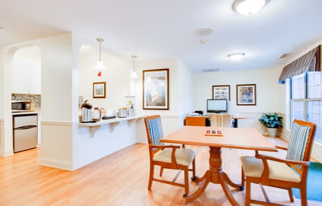 A bright and clean common area with a wooden table and four chairs with striped cushions. The room features light wood flooring, white walls, and two framed pictures on the walls. To the left, there is a kitchen area with white cabinets, a microwave, and a refrigerator. A counter with hanging pendant lights separates the kitchen from the seating area. A window with a valance lets in natural light, and a potted plant is placed near the window.