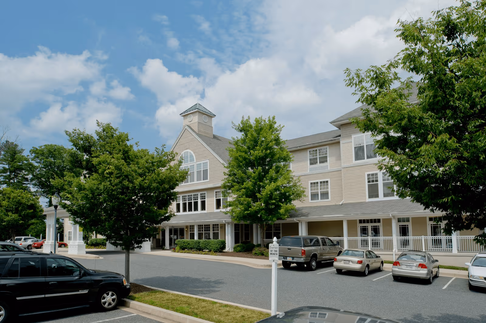 Exterior view of a multi-story senior living facility building with beige siding, multiple windows, and a small tower feature on the roof. There are several trees and parked cars in the parking lot in front of the building under a partly cloudy sky.