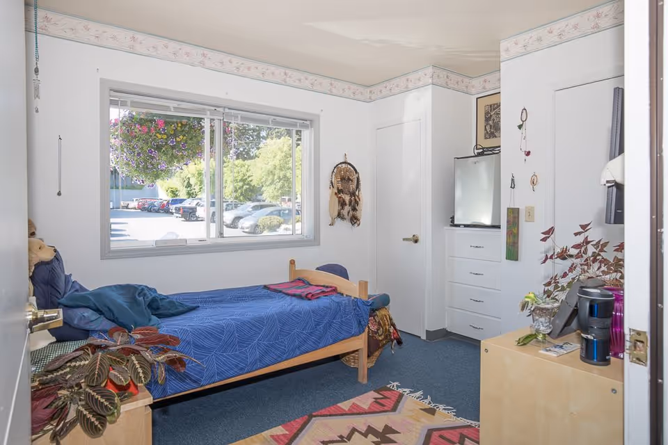 A small, cozy bedroom with a single wooden bed covered in a blue patterned bedspread and a folded red blanket at the foot. There is a large window with a view of parked cars and greenery outside. The room has white walls with a floral border near the ceiling, a dresser with a small refrigerator on top, and various decorative items including a dreamcatcher and plants. A colorful patterned rug is on the blue carpeted floor.