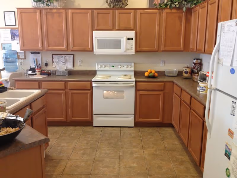A kitchen with wooden cabinets, a white electric stove with an oven, a white microwave above the stove, a white refrigerator on the right, and a countertop with various items including a bowl of oranges and kitchen utensils. The floor is tiled and there are plants on top of the cabinets.