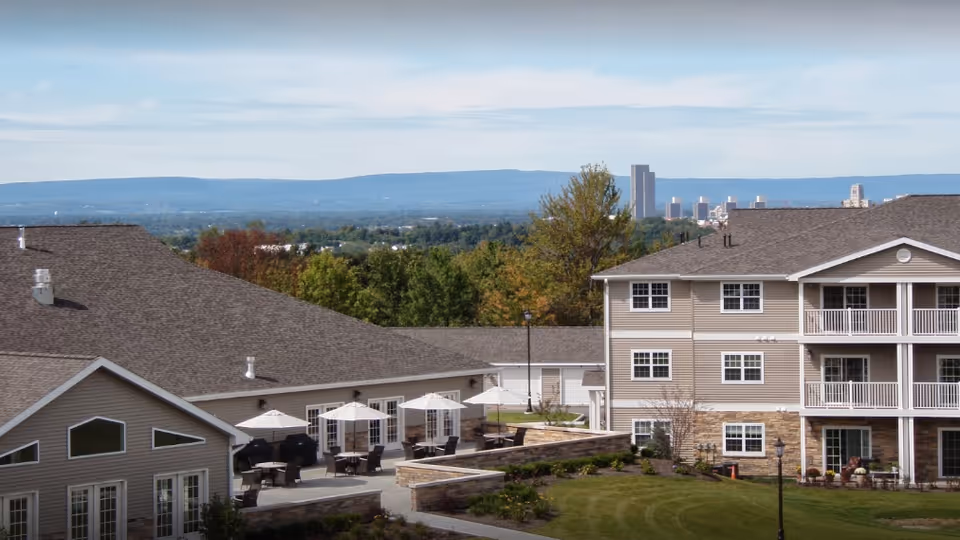 Exterior view of multi-story senior living buildings with balconies, patio umbrellas and landscaped lawn, with a distant city skyline and hills.
