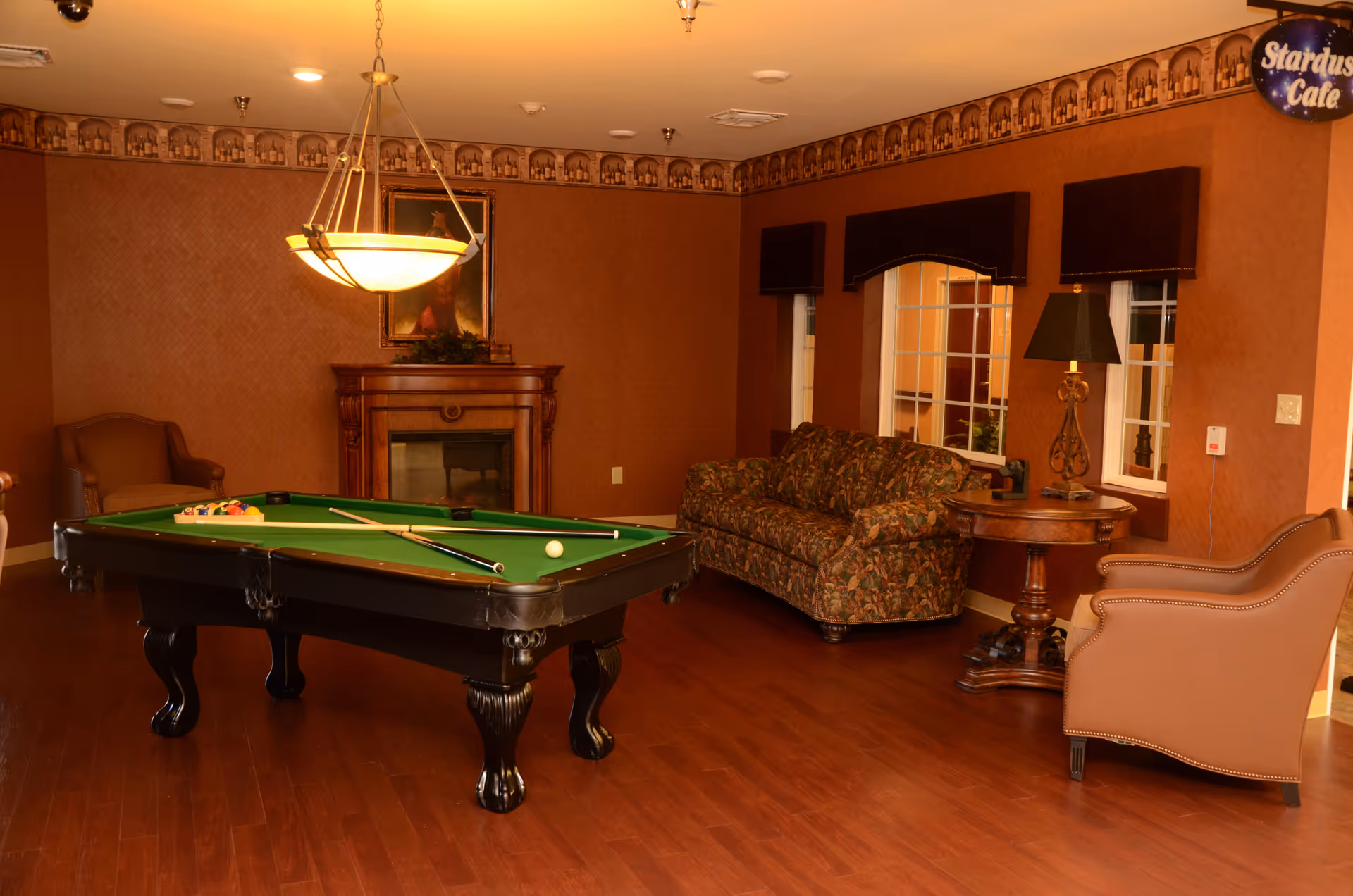 Interior lounge with a pool table, patterned sofa, armchairs, side table with lamp, and a fireplace.