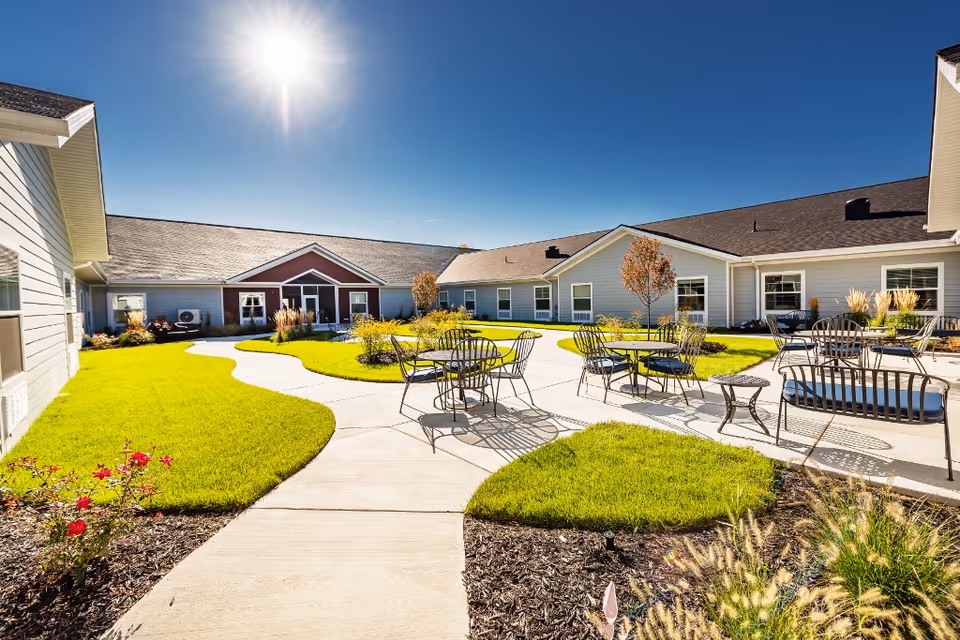 Sunny outdoor courtyard area at Provision Living at Fenton with green grass, paved walkways, metal tables and chairs, small trees, and surrounding single-story buildings under a clear blue sky.