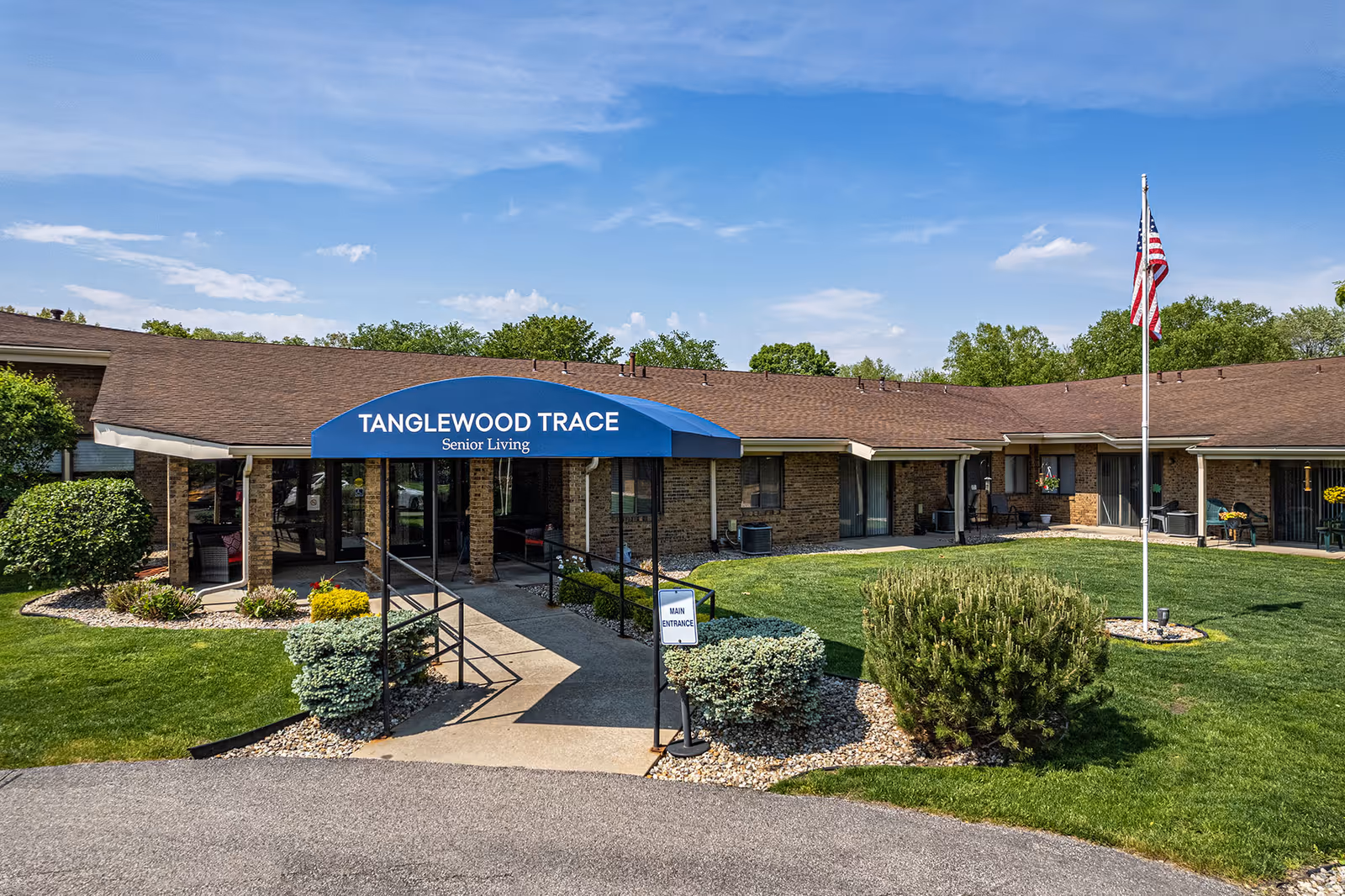 Exterior view of Tanglewood Trace Senior Living facility showing a single-story brick building with a brown roof, a blue awning over the main entrance, a paved walkway with handrails, manicured bushes, green lawn, and an American flag on a flagpole under a partly cloudy sky.