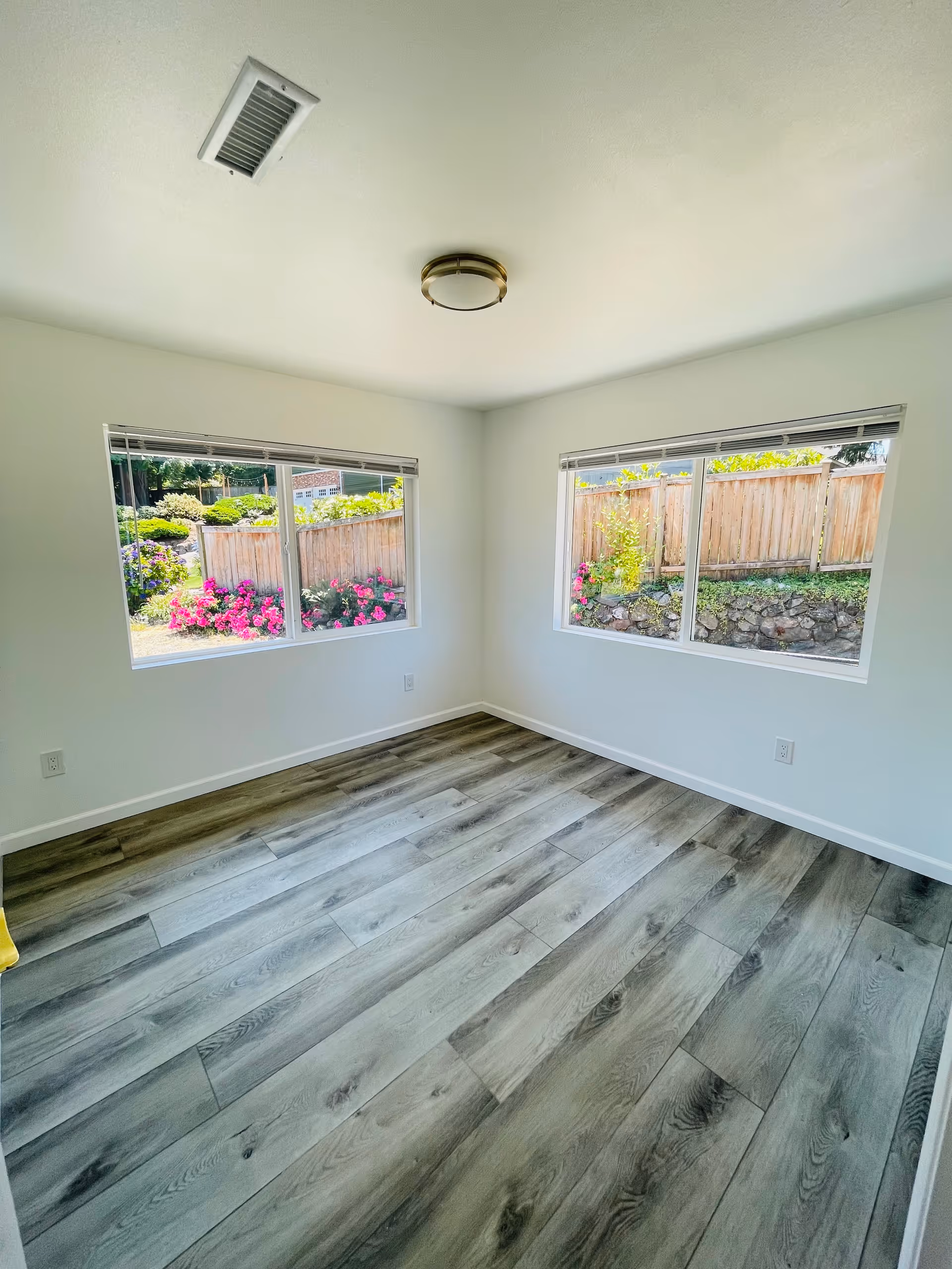 Empty room with light gray wood flooring, white walls, two large windows showing a garden with flowers and a wooden fence outside, and a ceiling light fixture.