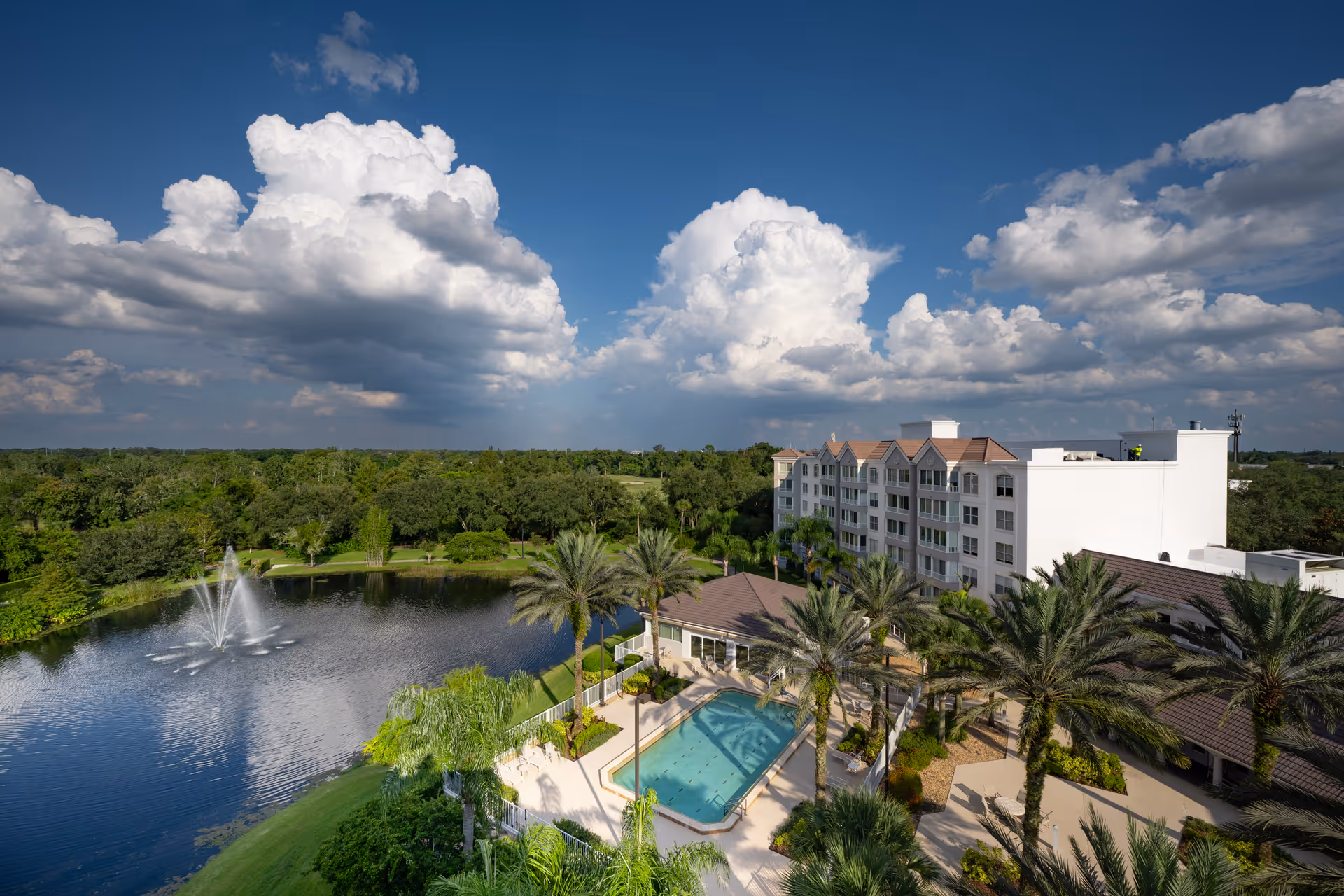 Aerial view of The Mayflower at Winter Park facility showing a large pond with a water fountain, a swimming pool surrounded by palm trees, and a multi-story residential building under a partly cloudy blue sky.