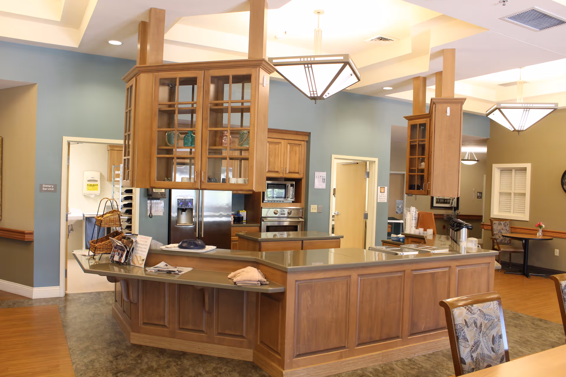 Interior view of a senior living facility kitchen and dining area with wooden cabinetry, a stainless steel refrigerator, built-in oven, and a countertop with various items. The walls are painted in blue and beige tones, and there are hanging light fixtures above the counter. Chairs with patterned upholstery are visible in the foreground and background.
