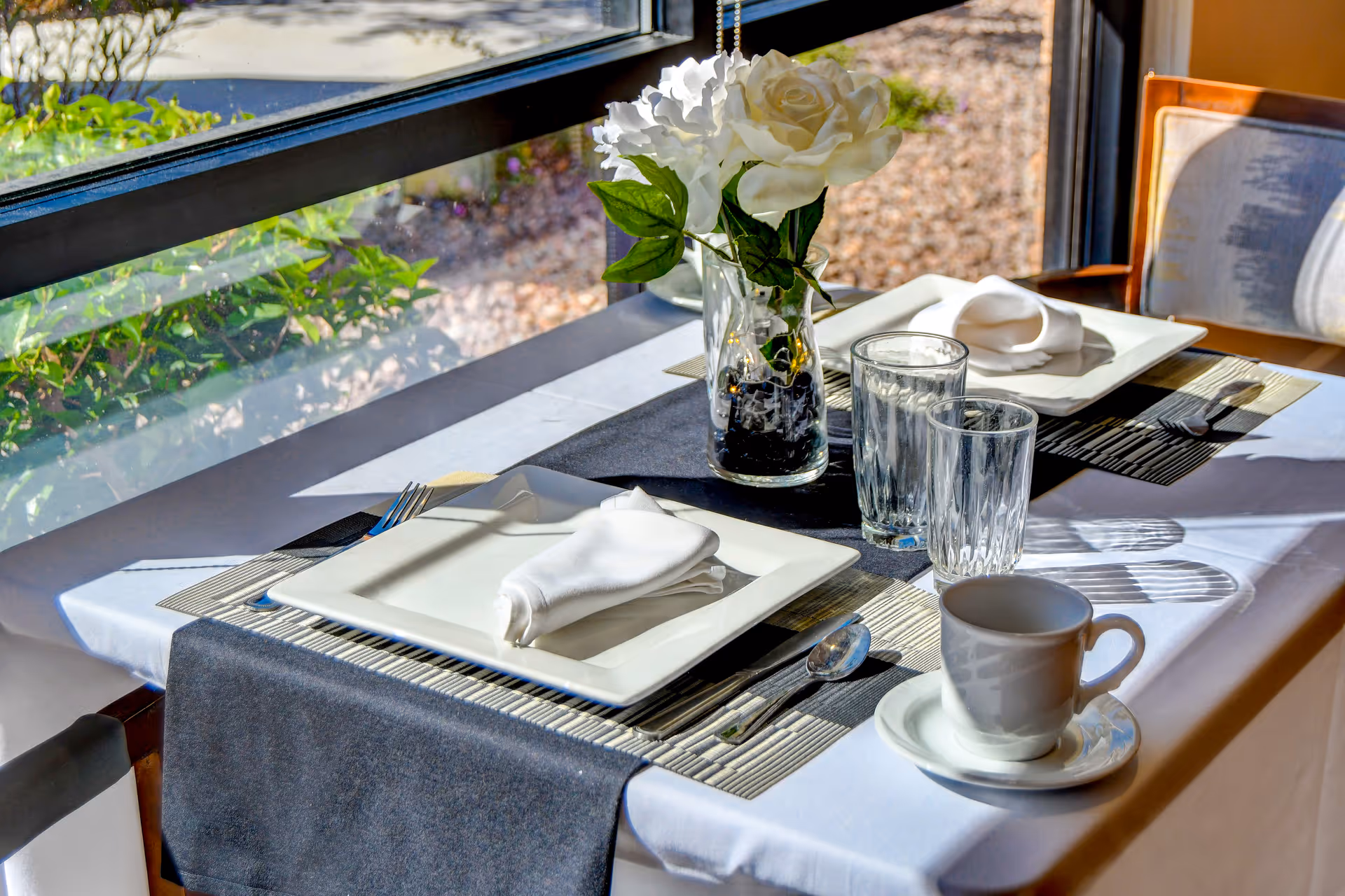 A neatly set dining table for two by a large window with sunlight streaming in. The table features white square plates with folded white napkins, clear drinking glasses, a white coffee cup on a saucer, silverware, and a vase with white flowers as a centerpiece.