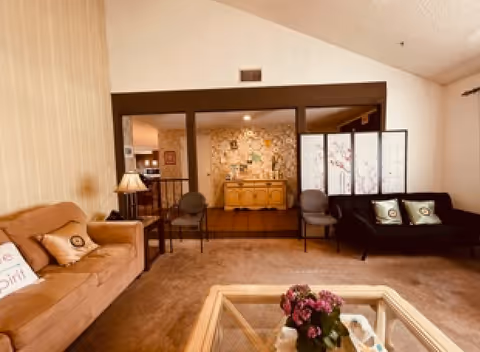 A cozy living room with a beige couch on the left, a black loveseat with two decorative pillows on the right, and a glass coffee table with a flower arrangement in the center. Behind the seating area is a wooden divider with a folding screen and two chairs. The background shows a sideboard against a wall with floral wallpaper and a warm, inviting ambiance.