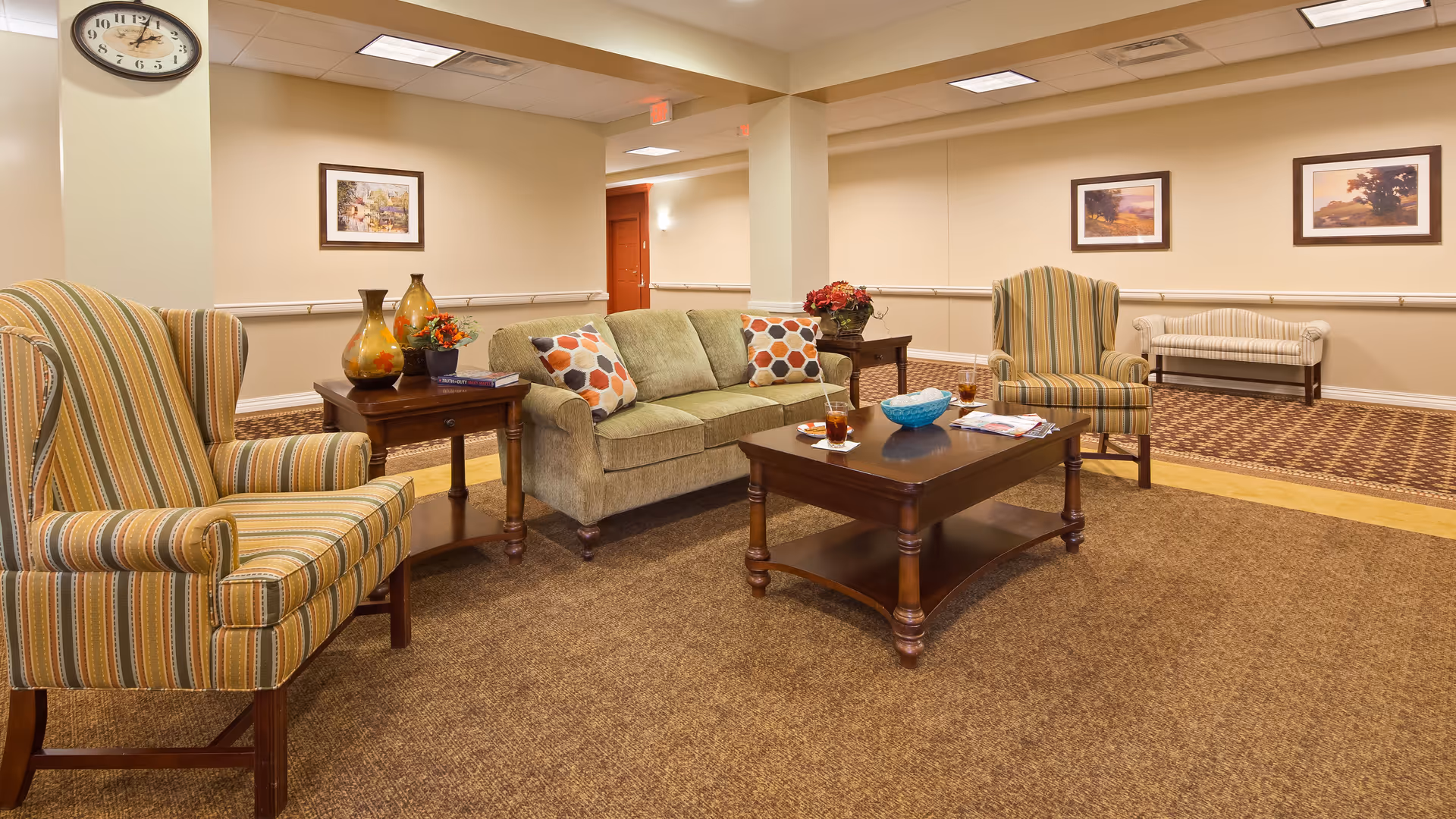 A cozy living room area in a senior living facility with a green sofa adorned with patterned cushions, two striped armchairs, a wooden coffee table with drinks and magazines, and side tables with decorative vases and flowers. The walls are beige with framed artwork, and there is a clock on one wall. The floor is carpeted with a mix of brown and patterned designs.
