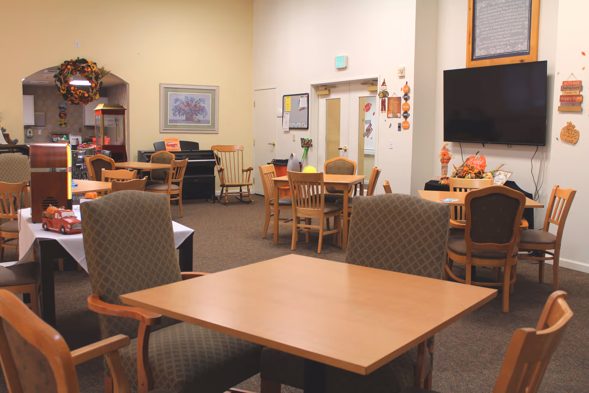 A senior living facility common area with multiple wooden tables and chairs arranged for dining or socializing. The room features a large flat-screen TV mounted on the wall, autumn-themed decorations, and a cozy atmosphere with carpeted floors. In the background, there is a kitchen area visible through an archway, and a piano against the wall.
