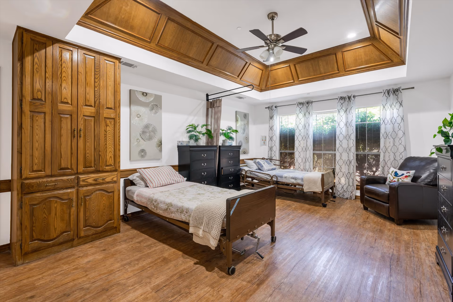A well-lit bedroom with two hospital-style beds, each with bedding and pillows. The room features wooden flooring, a large wooden wardrobe, two black dressers with plants on top, a brown leather armchair, and large windows with patterned curtains allowing natural light to enter. The ceiling has a wooden tray design with a ceiling fan and lights.