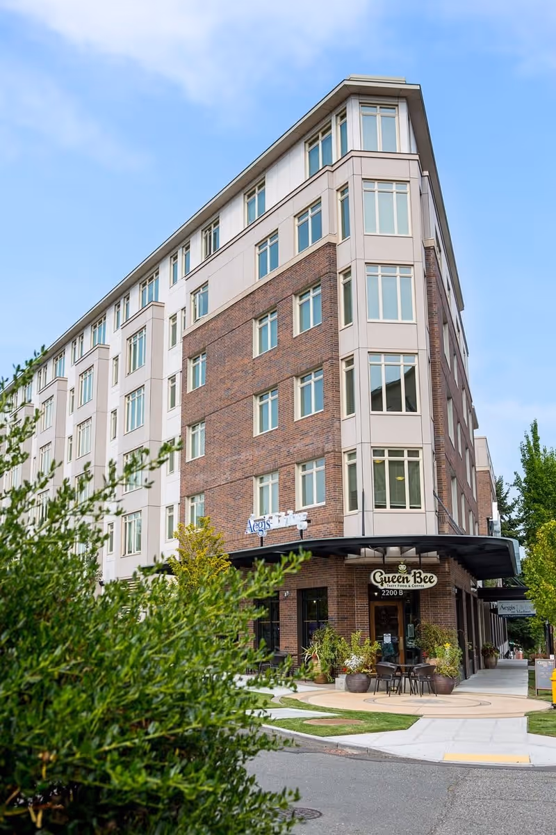 Exterior view of a multi-story senior living facility named Aegis Living Madison with a brick and beige facade. There is a small outdoor seating area with tables and chairs in front of a cafe called Queen Bee. Green shrubs and trees surround the building under a partly cloudy blue sky.