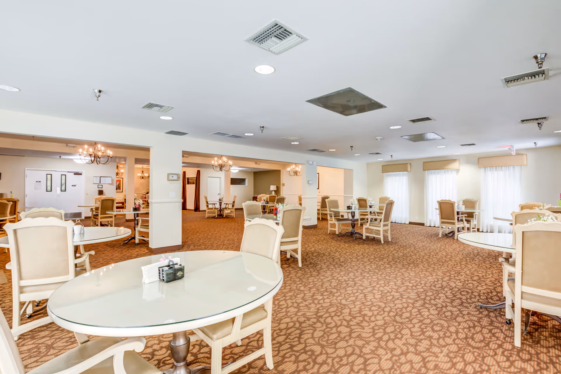 A spacious dining room in a senior living facility with multiple round tables and beige cushioned chairs arranged neatly. The room has carpeted flooring with a patterned design, white walls, and several windows with sheer curtains allowing natural light to enter. Chandeliers and ceiling lights provide additional illumination.