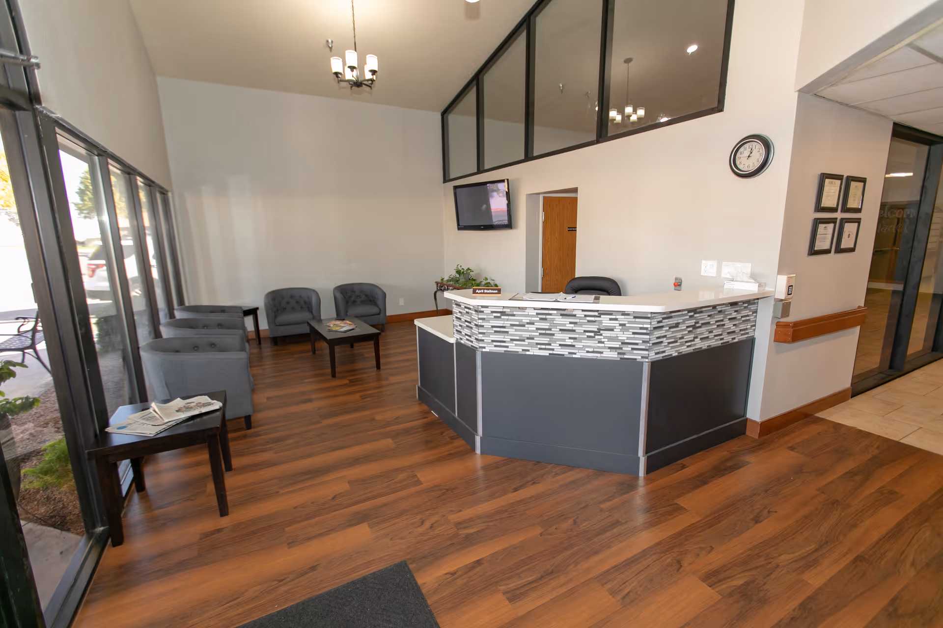 Reception area of a senior living facility with a modern front desk featuring a tiled front panel, a clock on the wall, a flat screen TV mounted above a doorway, and a seating area with gray armchairs and a coffee table. Large windows let in natural light and show a glimpse of the outdoor area.