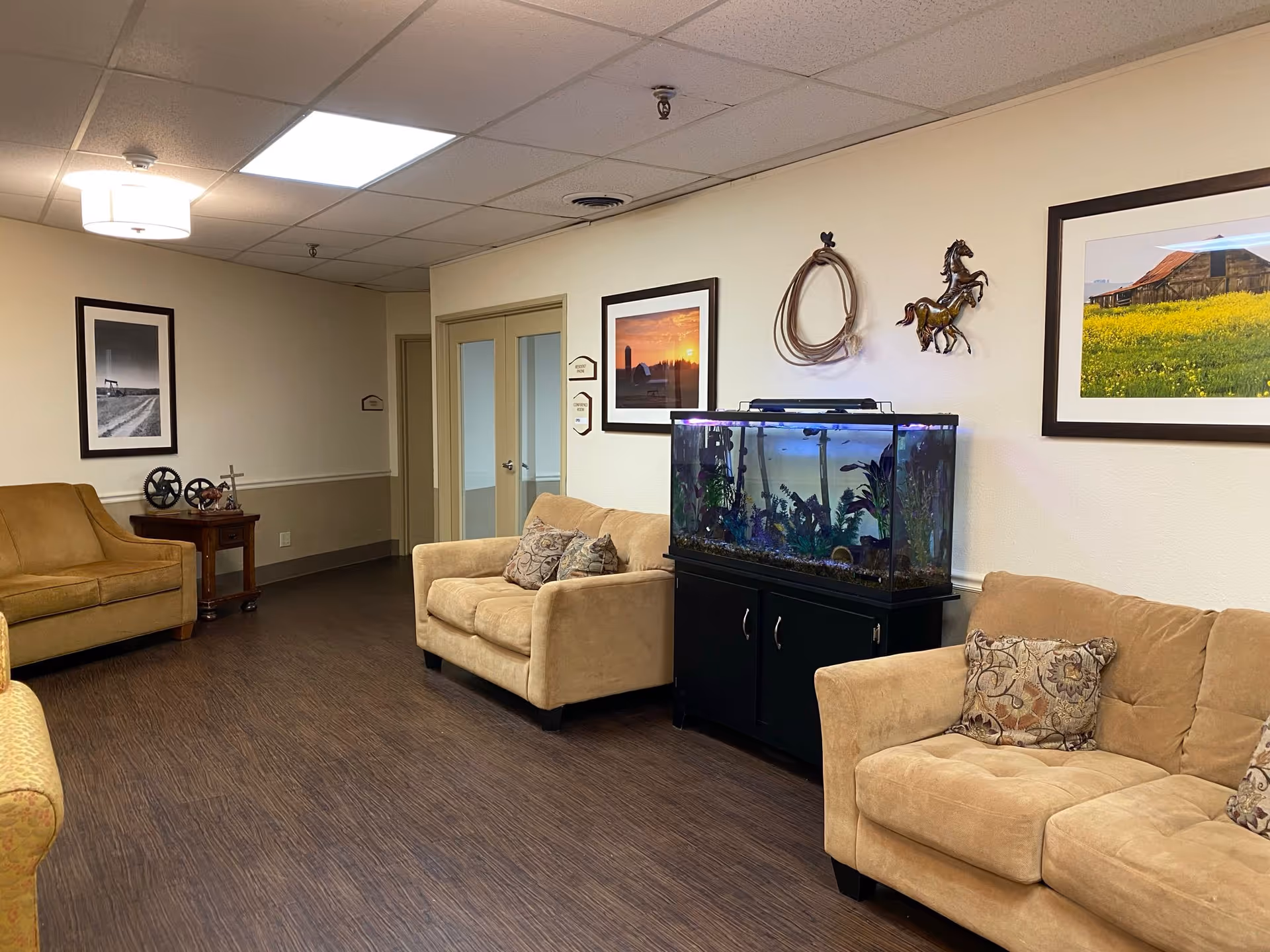 A cozy seating area in a senior living facility with beige sofas and armchairs arranged around a large fish tank on a black cabinet. The walls are decorated with framed pictures of rural scenes and metal horse sculptures. The floor has dark wood-like flooring and the ceiling has recessed lighting and a ceiling light fixture.