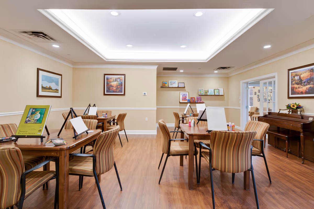 A well-lit activity room with wooden floors and beige walls, featuring several tables with striped chairs arranged around them. Each table has easels with blank canvases and art supplies, suggesting a painting or art class setup. The room is decorated with framed landscape paintings on the walls and a piano with a flower arrangement on top in the corner.