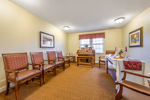 A small room with beige walls and carpeted floor featuring six wooden chairs with red cushioned seats arranged along the walls. There is a wooden organ against the far wall beneath a window with a red valance. A table covered with a white cloth and red runner holds decorative items including candles and flowers. Two framed paintings hang on the walls.