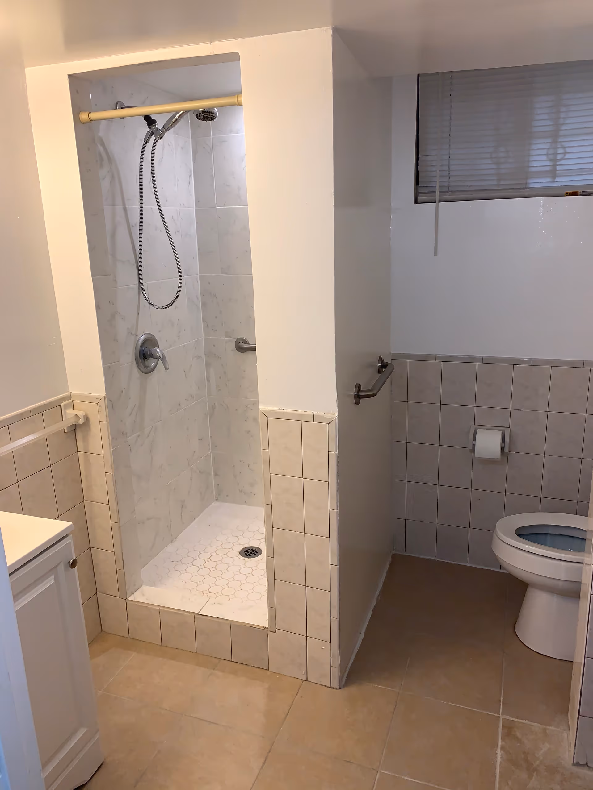 A bathroom with a walk-in shower featuring a handheld showerhead and tiled walls. There is a toilet with a grab bar on the adjacent wall and a small window with blinds above it. The floor is tiled, and part of a white vanity cabinet is visible on the left side.