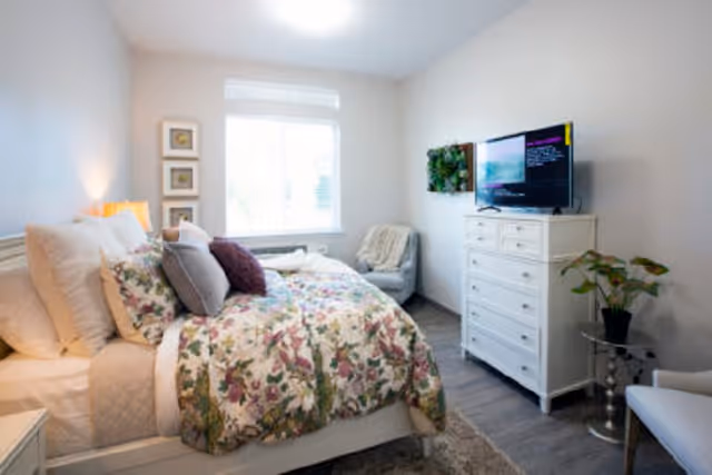 A cozy bedroom with a bed covered in floral patterned bedding and multiple pillows. There is a window letting in natural light, a white dresser with a TV on top, a small plant on a side table, and a comfortable chair with a blanket draped over it.