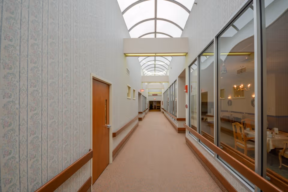 A long, well-lit hallway in a senior living facility with patterned wallpaper, wooden doors on the left, and large windows on the right showing a dining area with tables and chairs. The ceiling has a series of arched skylights allowing natural light to fill the corridor.