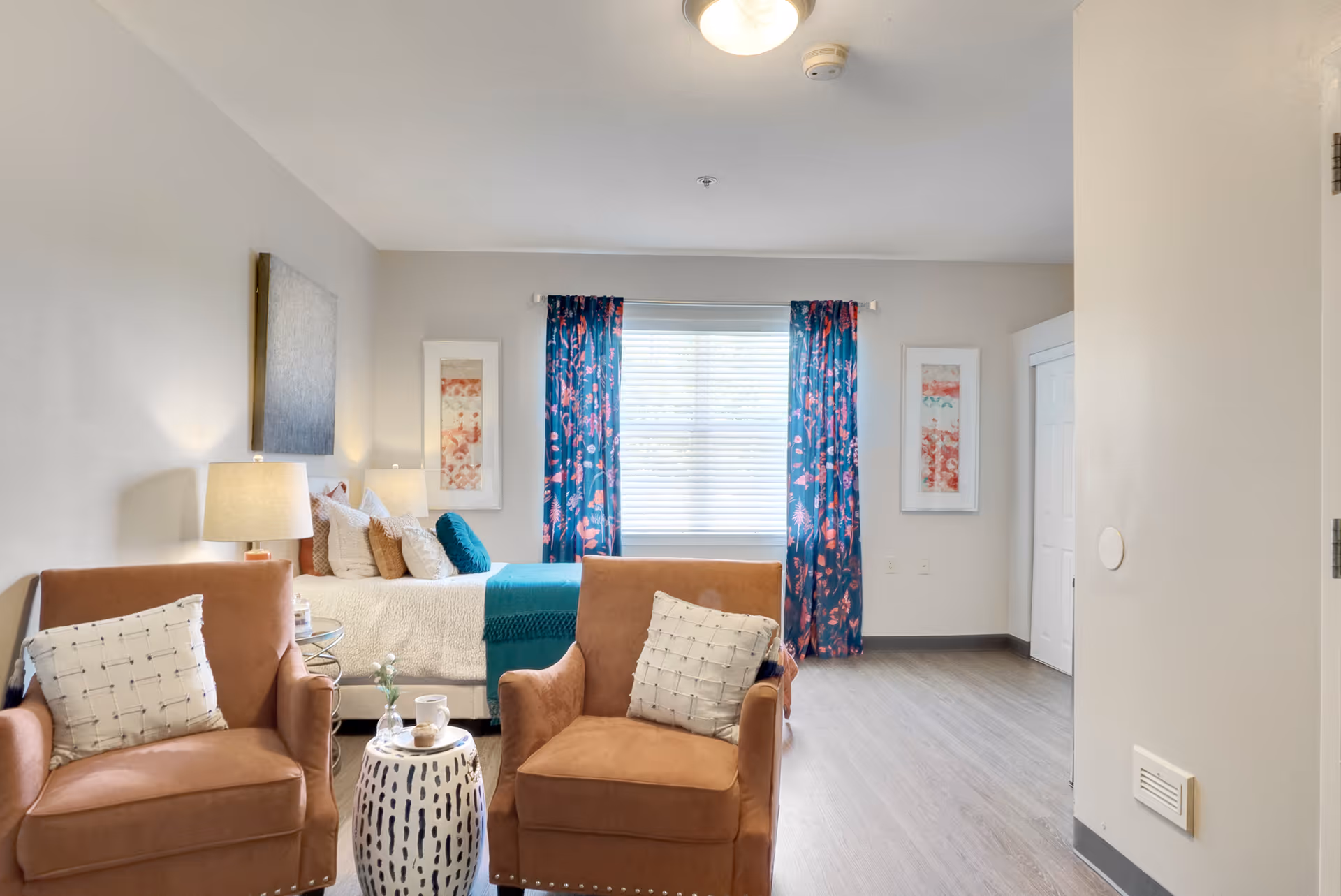 A bright and cozy bedroom featuring a bed with white bedding and teal accents, two brown armchairs with white pillows, a small white side table with a cup and vase, blue floral curtains on the window, and framed artwork on the walls.