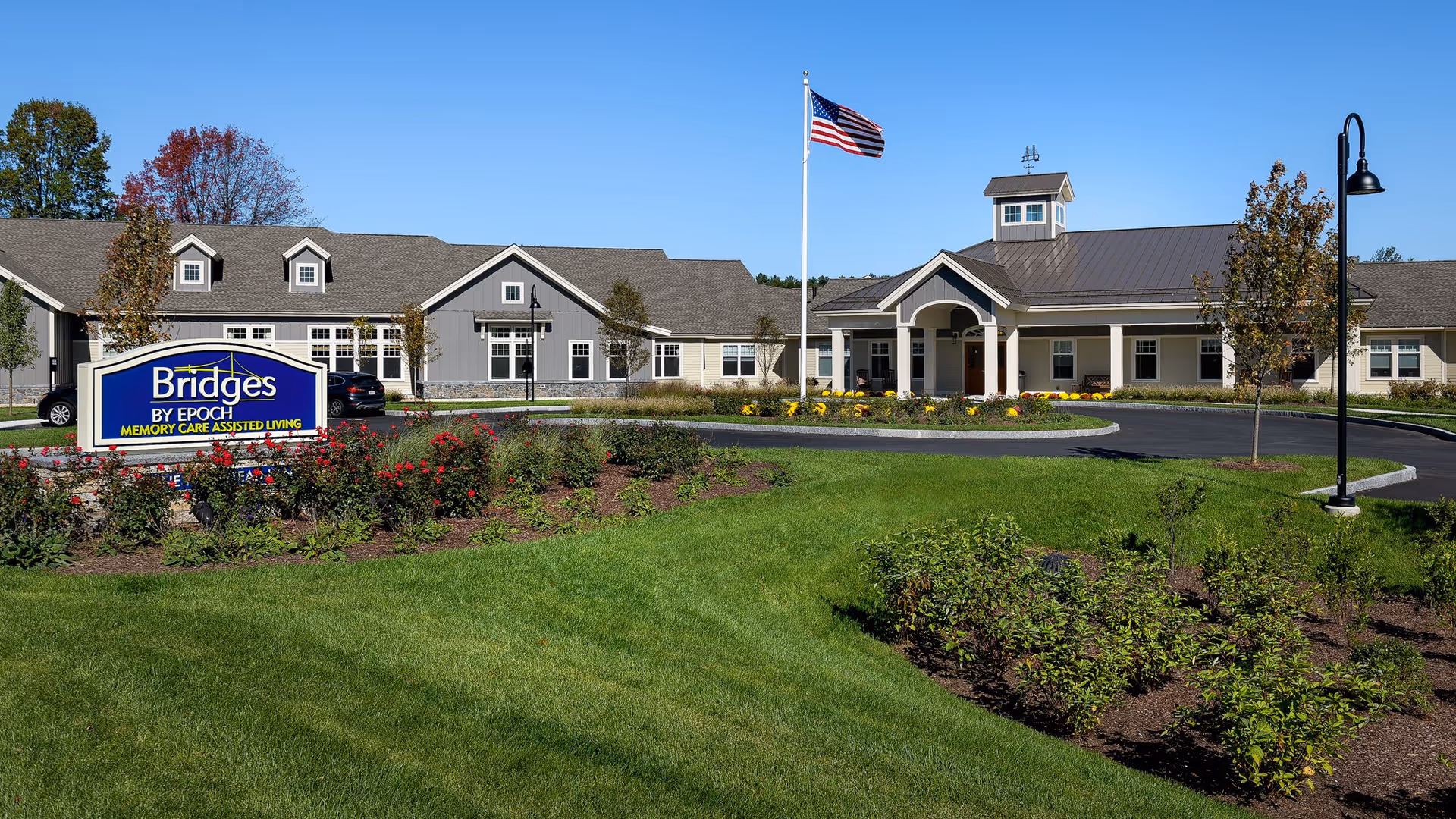 Exterior view of Bridges by EPOCH at Sudbury, a memory care assisted living facility, showing a single-story building with a gray roof and beige walls, a circular driveway with an American flag in the center, landscaped gardens with green grass, bushes, and flowers, and a blue sign with the facility's name in front.