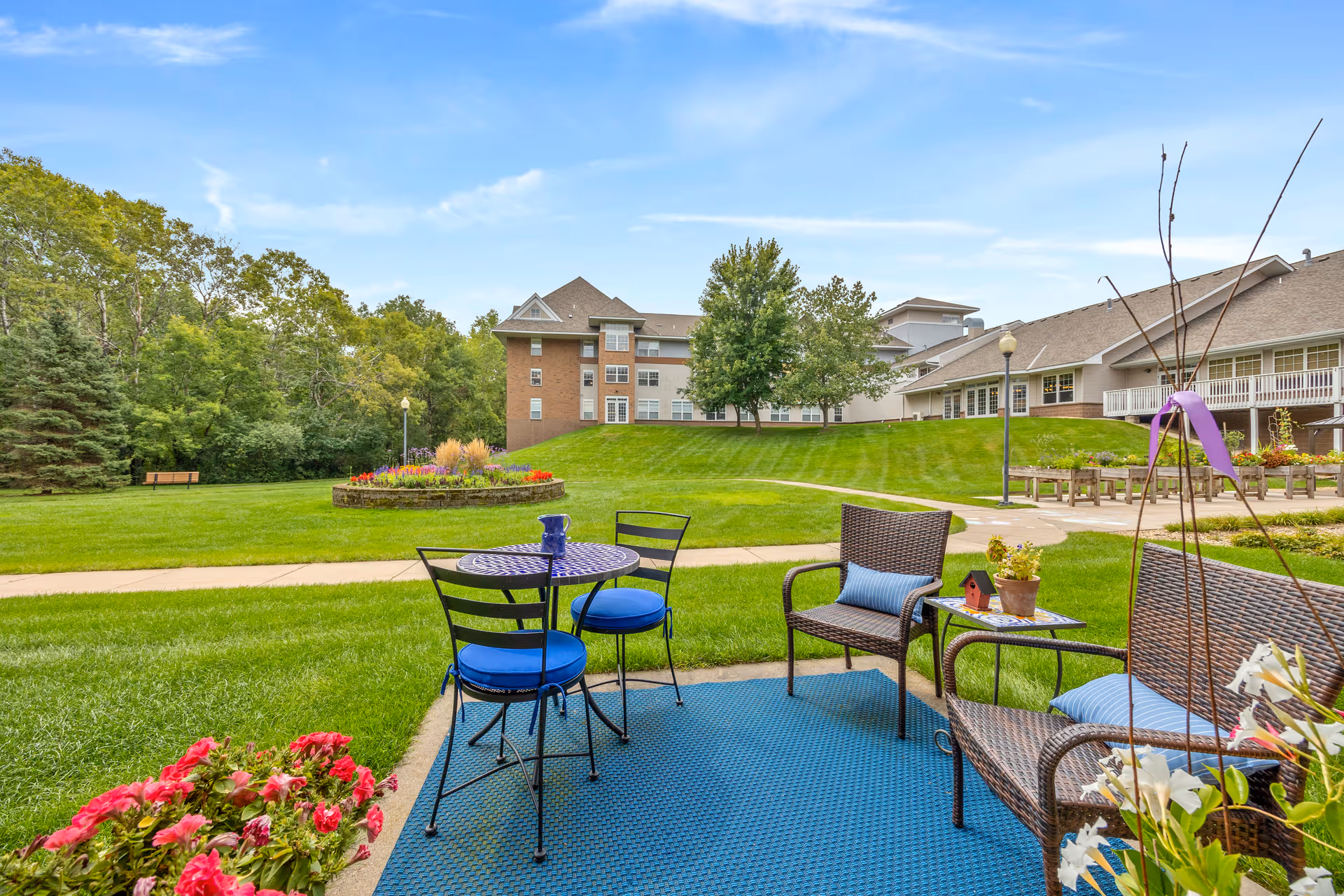 Outdoor seating area with a small round table and two chairs with blue cushions, and a wicker loveseat and chair with blue pillows on a blue outdoor rug. The area overlooks a well-maintained lawn with a circular flower bed, trees, and a multi-story building in the background under a blue sky.