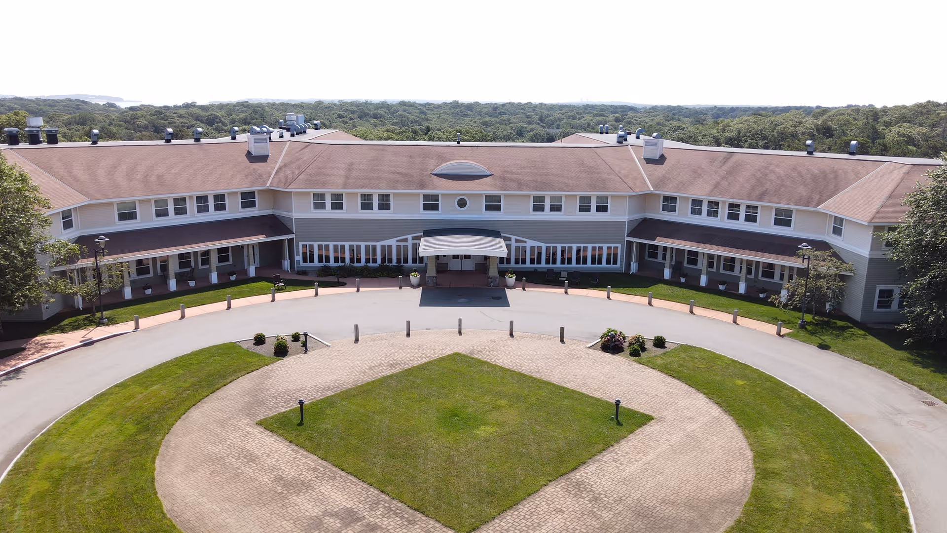 Aerial view of a large senior living facility building with a circular driveway and a landscaped grassy area in front. The building has a brown roof, multiple windows, and a covered entrance. Trees and forested area are visible in the background.