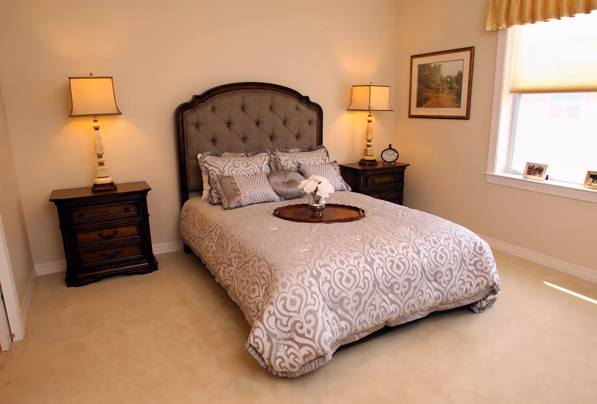 A neatly made bed with a tufted headboard in a bedroom. The bed has a patterned beige and white comforter with multiple decorative pillows. There are two wooden nightstands on either side of the bed, each with a lamp. A small round tray with white flowers is placed on the bed. A framed picture hangs on the wall to the right, and a window with a beige shade lets in natural light.