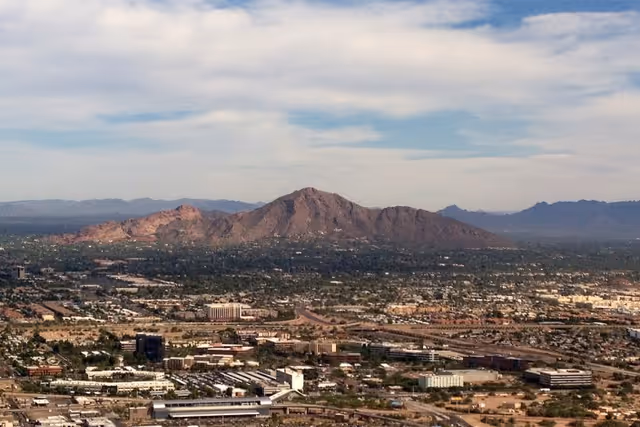 A panoramic aerial view of a city with a prominent mountain range in the background under a partly cloudy sky.