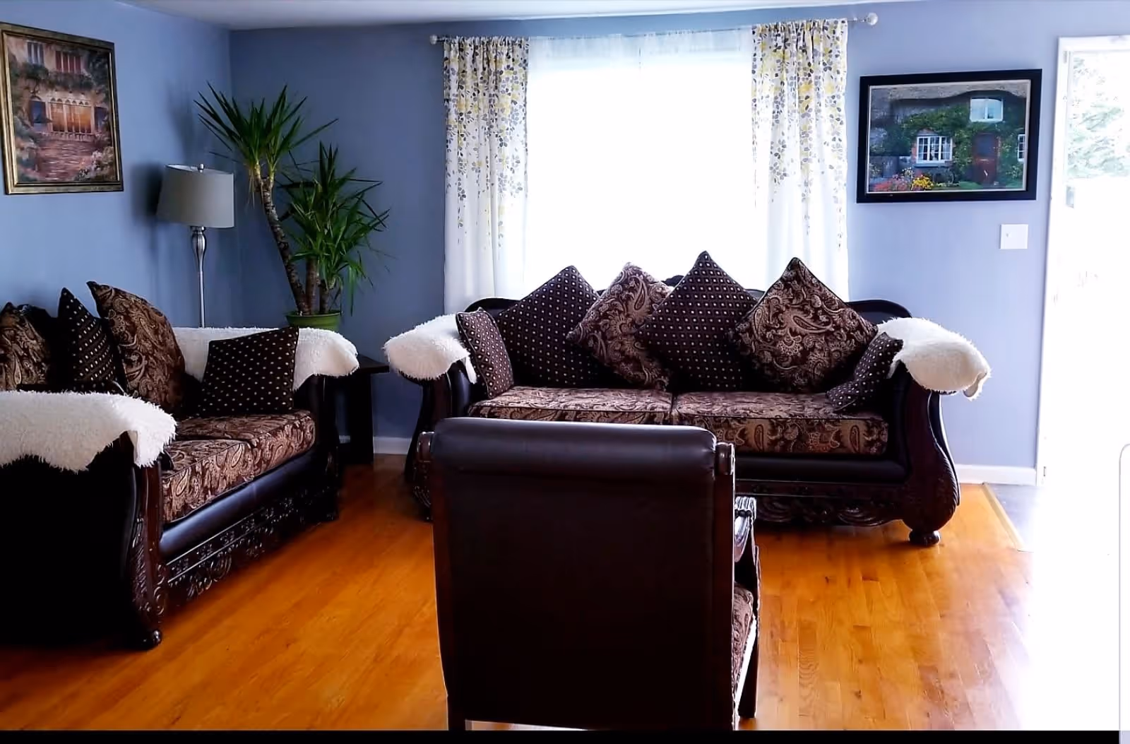 A cozy living room with two dark wood sofas and a matching armchair, all upholstered with patterned cushions and white throws. The room has light blue walls, wooden flooring, a tall green plant in the corner, a floor lamp, and two framed paintings on the walls. White curtains with a floral pattern cover a large window letting in natural light.