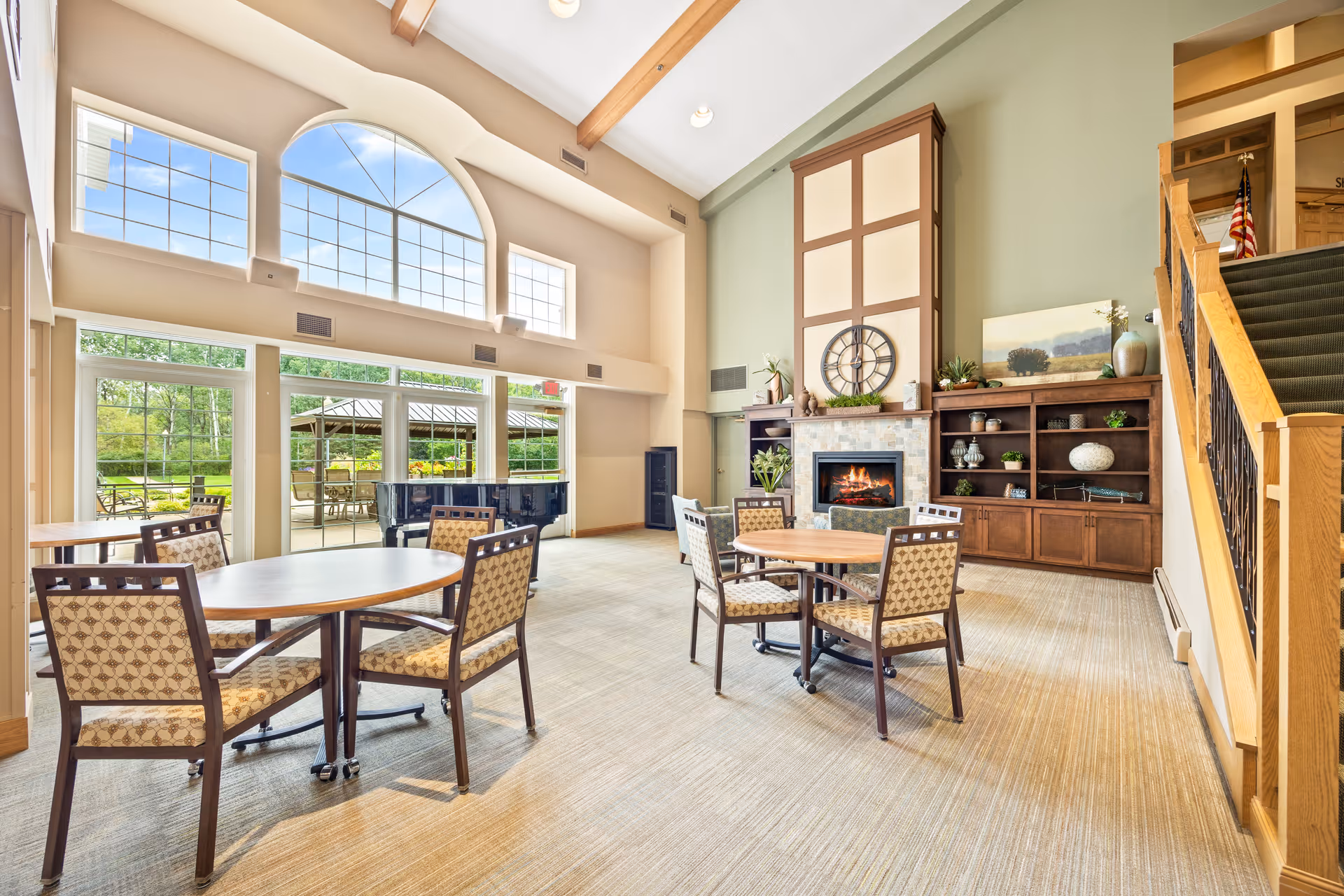 Bright communal living area with round tables and chairs, a fireplace and built-in shelving, large arched windows, and a piano.