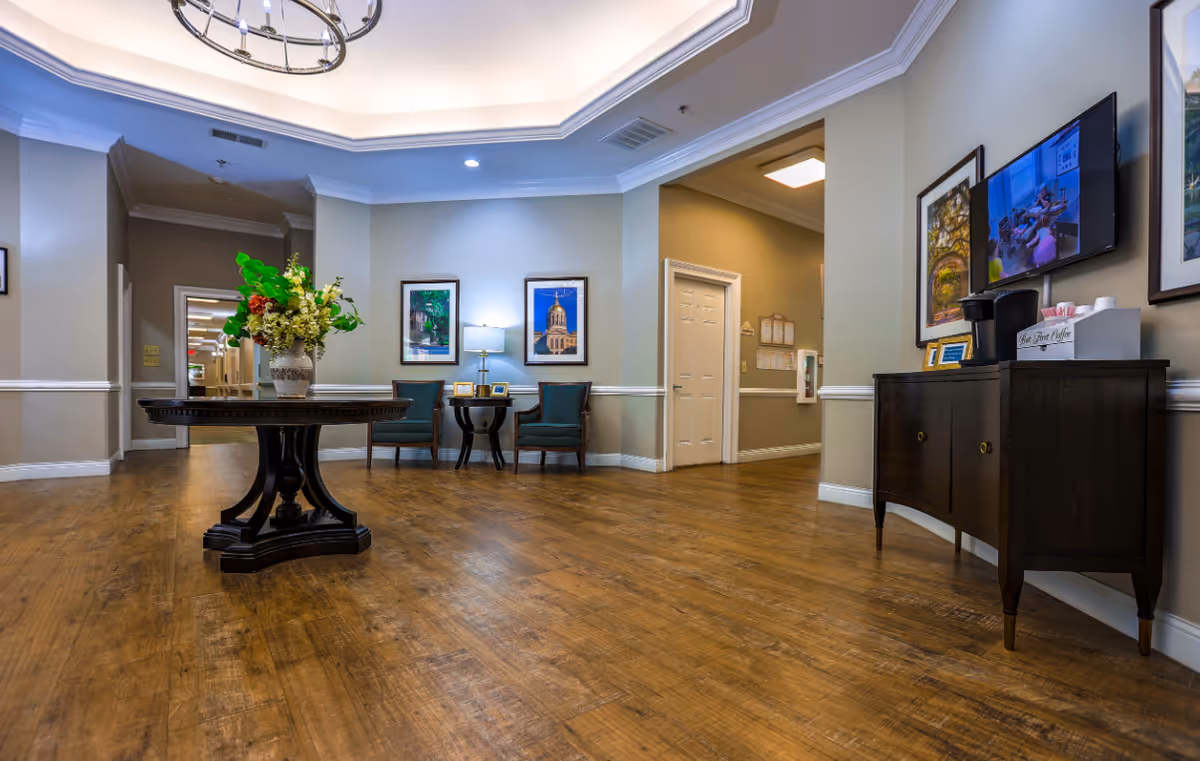 A spacious, well-lit interior room with wooden flooring and light-colored walls. In the center is a round wooden table with a large floral arrangement. Against the far wall are two green upholstered chairs with a small table and lamp between them. On the right side, a dark wooden cabinet holds a coffee maker and supplies, with a flat-screen TV mounted above it. Several framed pictures hang on the walls, and a chandelier is visible on the ceiling.