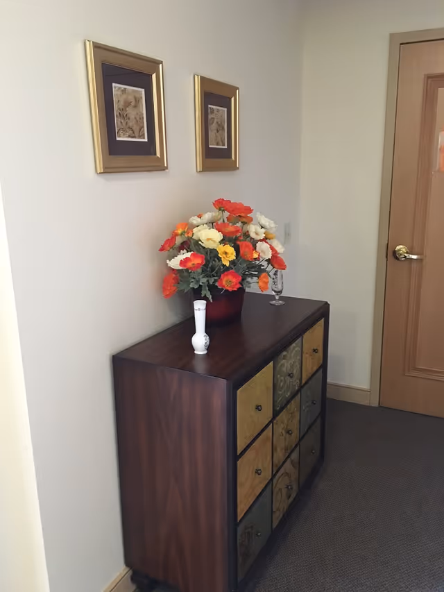 A wooden dresser with colorful artificial flowers and small vases beneath two framed pictures in a hallway next to a closed door.