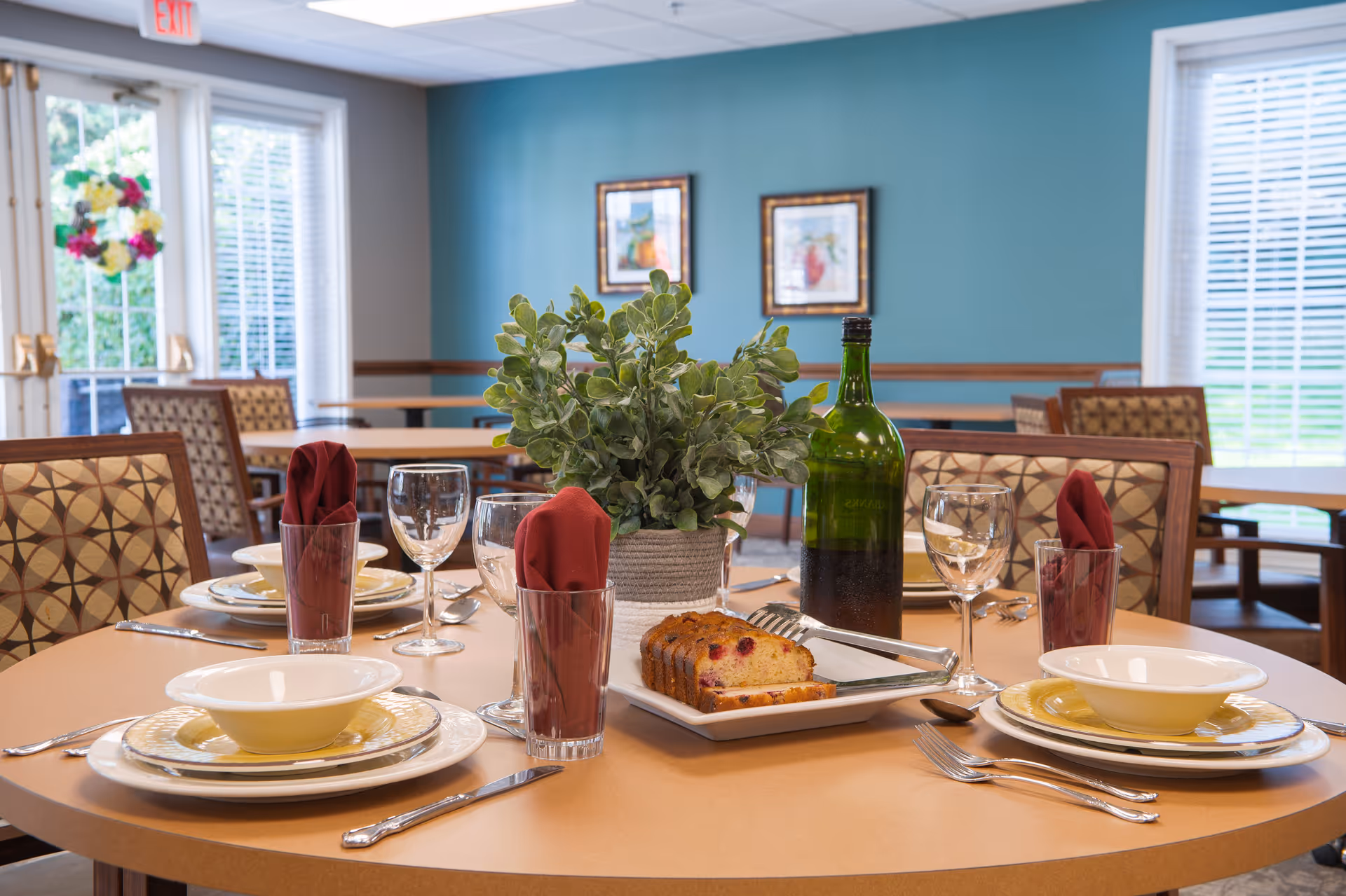 A round dining table set for four with plates, bowls, silverware, wine glasses, and glasses with red cloth napkins. A green bottle and a plate with sliced cake are in the center, along with a potted plant. The room has patterned chairs, teal walls with framed pictures, and large windows letting in natural light.