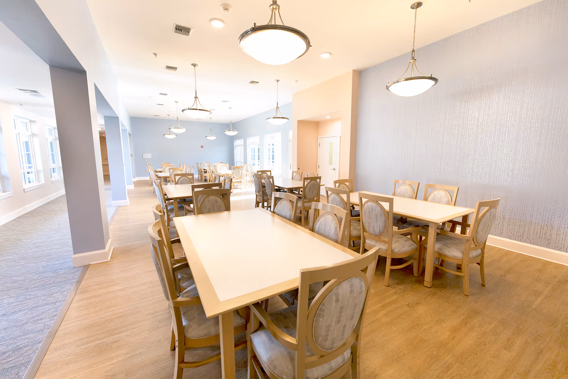 A bright dining room with multiple rectangular tables and wooden chairs with cushioned seats and backs. The room has light-colored walls, large windows letting in natural light, and several hanging ceiling lights. The floor is a combination of wood and carpet.