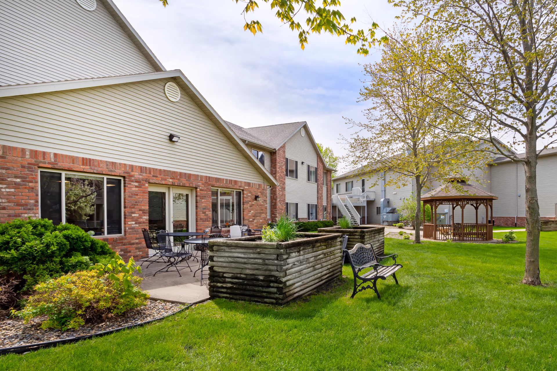 Outdoor view of Mankato Lodge Senior Living showing a grassy courtyard with trees, a wooden gazebo, a bench, raised garden beds, and patio tables and chairs next to a brick and siding building under a partly cloudy sky.