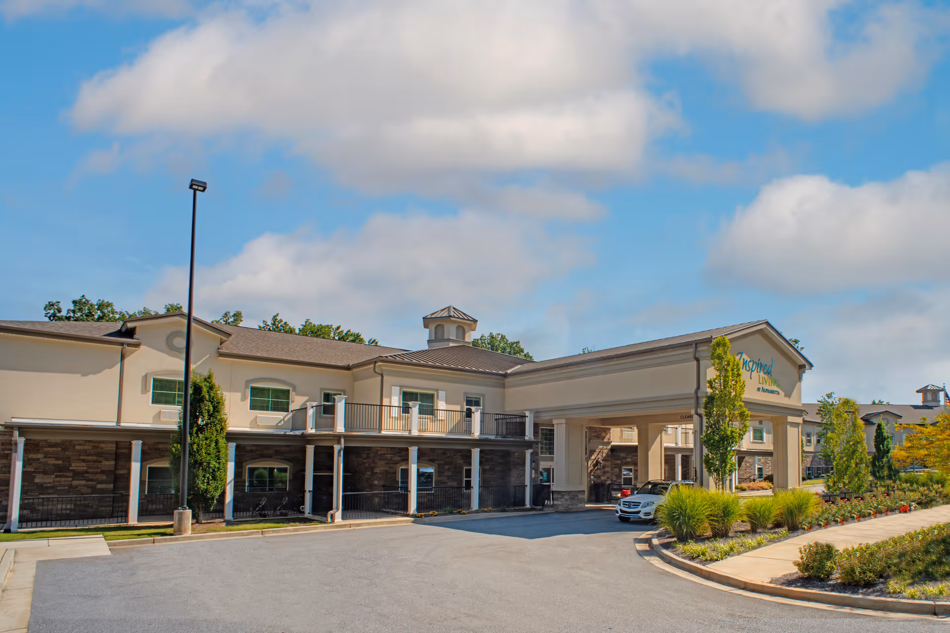 Exterior view of Inspired Living Alpharetta facility showing a two-story building with a covered entrance, landscaped greenery, and a parked car under a partly cloudy sky.