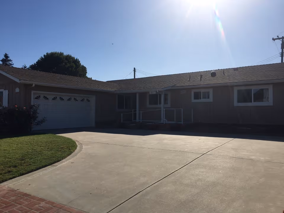 Single-story building front with an attached two-car garage, wheelchair ramp, small lawn, and a wide paved driveway under a sunny sky.