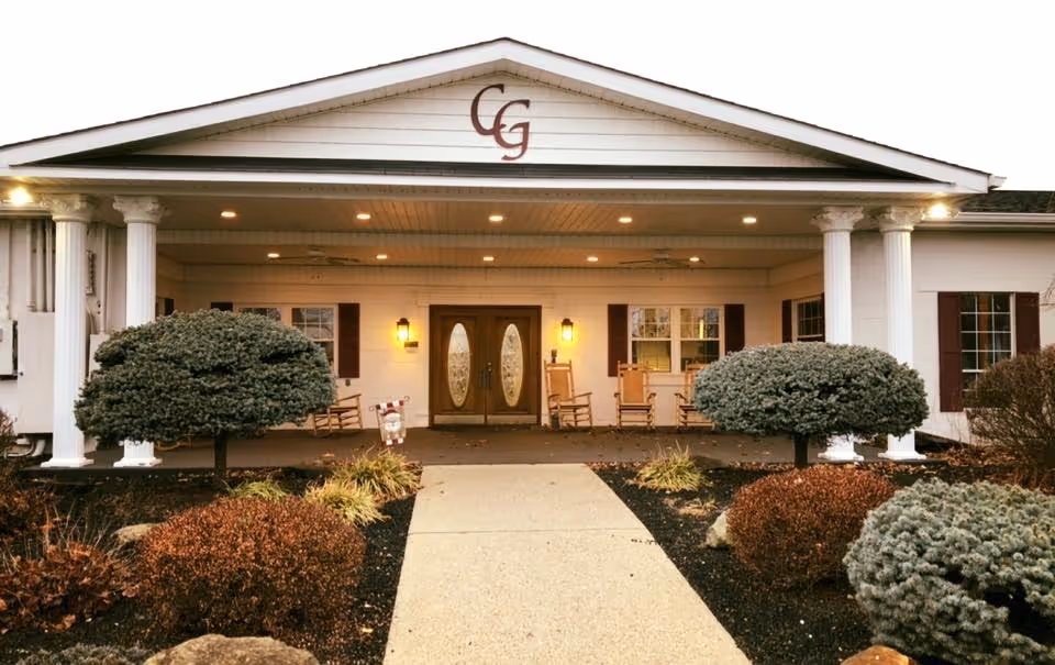 Front exterior view of Cedar Grove facility showing a covered entrance with white columns, double glass doors, rocking chairs on the porch, and neatly trimmed bushes and landscaping along the walkway.