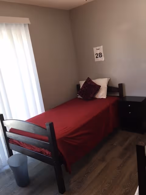 A small bedroom with a single bed covered in a red blanket and two pillows, one white and one dark red. The bed is positioned against a gray wall with a paper sign labeled '2B' taped above the headboard. Next to the bed is a black nightstand with two drawers. A window with white vertical blinds is on the left side, allowing natural light into the room. The floor is wooden, and there is a small gray trash bin near the foot of the bed.