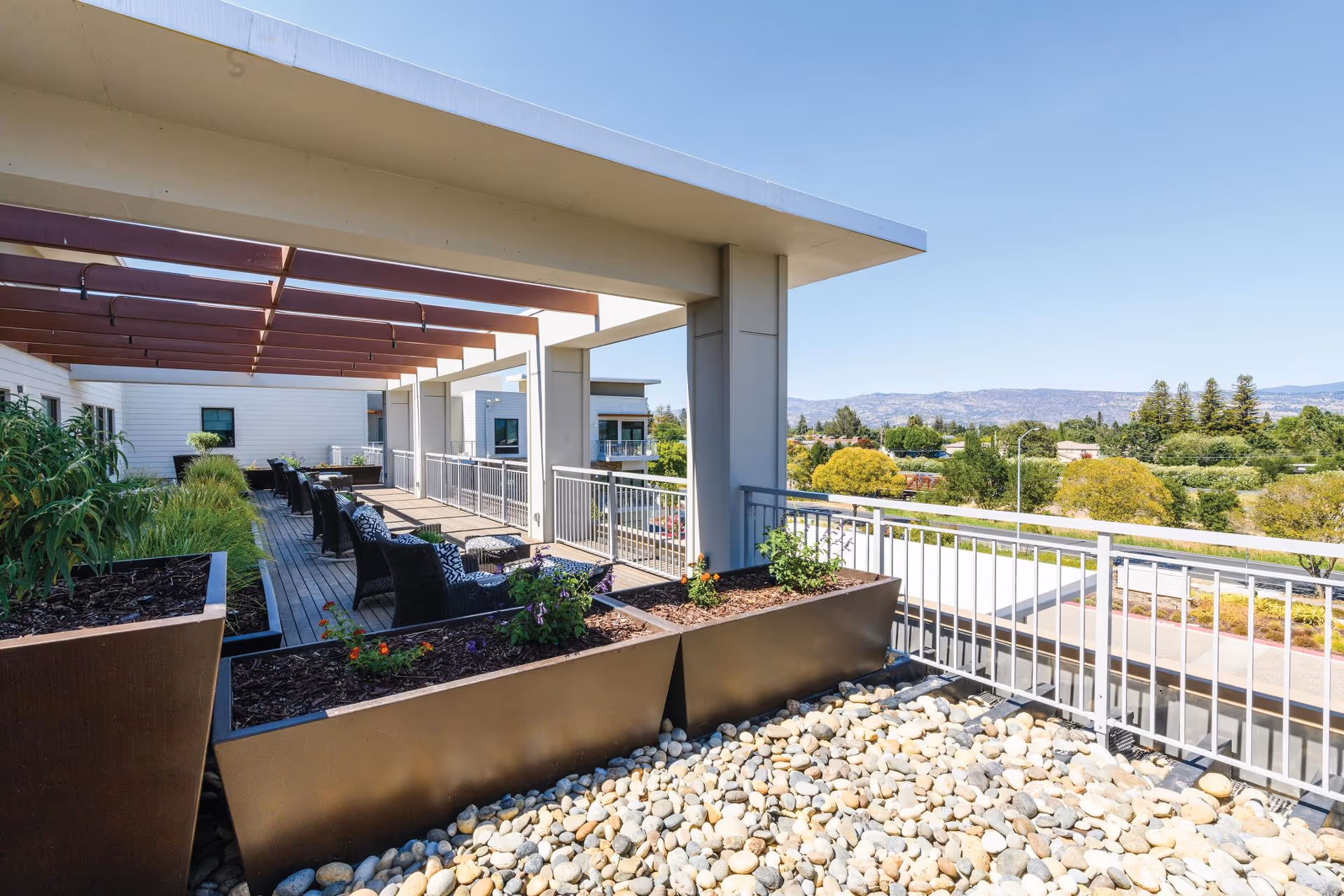 Outdoor patio area at The Watermark at Napa Valley featuring a wooden deck with black wicker chairs and small tables, large planters with greenery and flowers, a pergola overhead, and a view of trees and hills in the distance under a clear blue sky.