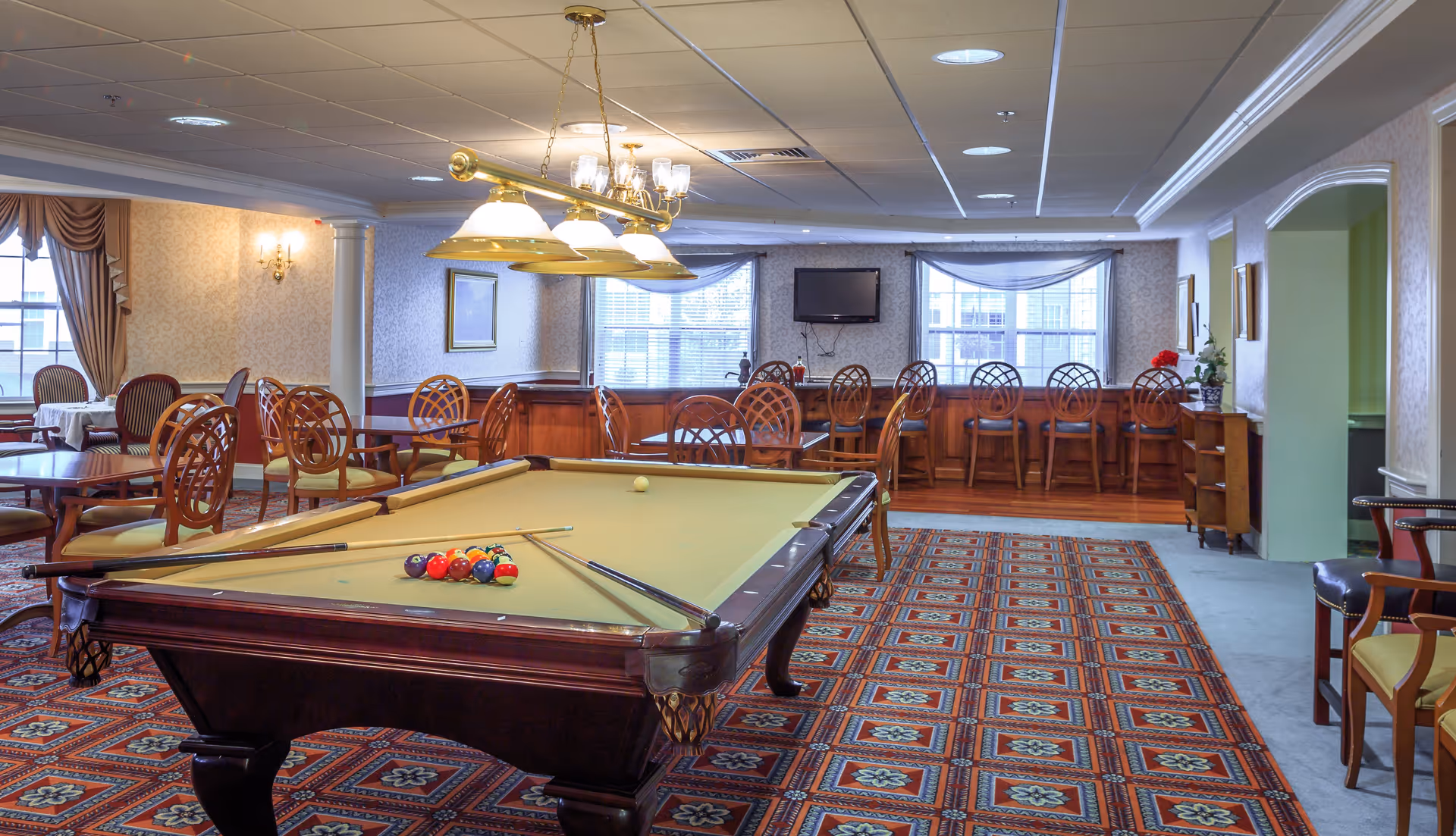 A spacious recreational room with a pool table in the center, surrounded by wooden chairs and tables. The room has patterned carpet flooring, large windows with curtains, a wall-mounted TV, and a row of high chairs along a counter. The lighting includes a chandelier above the pool table and wall sconces.